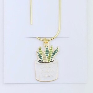 May include: A gold-colored bookmark with a dangling charm featuring a potted succulent. The charm has green and white details, and the pot reads "crazy plant lady." The bookmark is attached to a white card with the text "Duchess Gems Studio."