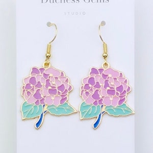 May include: A pair of gold-tone earrings featuring a pink and purple hydrangea flower design with green leaves and blue stems. Duchess Gems Studio.