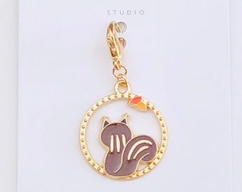 May include: Gold charm featuring a brown squirrel with a white tail inside a gold circle with a small red and orange leaf. The charm is attached to a gold chain.