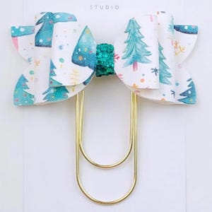 May include: A decorative paper clip featuring a Christmas tree bow. The bow is white with a pattern of blue and green Christmas trees and snowflakes. The center of the bow is a teal glitter. The paper clip is gold and the card reads "Duchess Gems STUDIO".