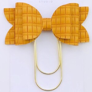 May include: A gold paper clip with a large, mustard yellow bow made of faux leather with a textured pattern. The bow is attached to the paper clip. The text "Duchess Gems Studio" is printed on a white card behind the bow.