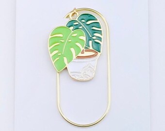 May include: Gold metal bookmark with a green and white enamel plant design. The bookmark features a potted plant with two leaves, one green and one light green. The text "Duchess Gems Studio" is printed on the white background.