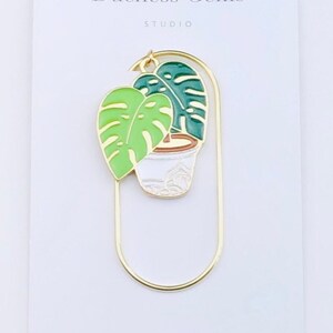 May include: Gold metal bookmark with a green and white enamel plant design. The bookmark features a potted plant with two leaves, one green and one light green. The text "Duchess Gems Studio" is printed on the white background.