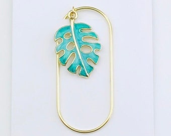 May include: Gold bookmark with a teal green monstera leaf charm. The bookmark is shaped like a long oval. Duchess Gems Studio is printed on the card behind the bookmark.
