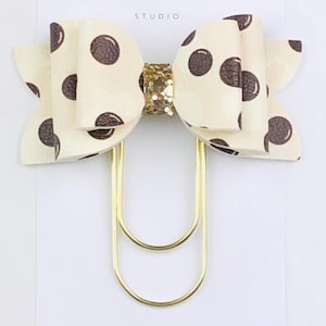 May include: A large paperclip featuring a cream-colored bow with brown polka dots and a gold glitter center. The bow is attached to a gold-tone metal paperclip and is mounted on a white display card with the text Duchess Gems STUDIO.