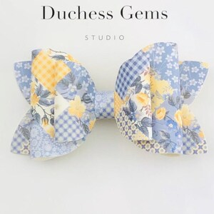 Könnte beinhalten: Eine Haarspange mit Blumenmuster in Blau-, Gelb- und Weißtönen, mit dem Text "Duchess Gems STUDIO" auf weißem Hintergrund. Die Schleife hat ein Patchwork-Design mit verschiedenen Blumen- und geometrischen Mustern.