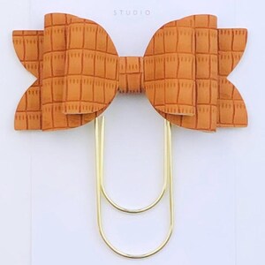 May include: A gold paper clip with a large, orange, faux leather bow with a checkered pattern. The bow is attached to the paper clip. The paper clip and bow are on a white card with the text "Duchess Gems STUDIO" printed on it.