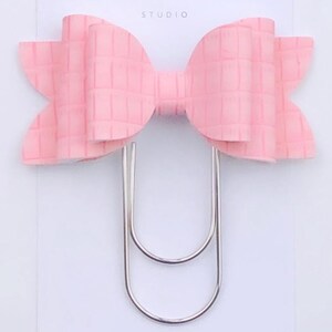 May include: A pink bow with a checkered pattern is attached to a silver double-pronged binder clip. The clip is on a white card with the text "Duchess Gems Studio".