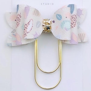 May include: A white bow with a colourful abstract print and a gold glitter centre is attached to a gold double-prong paper clip. The bow is labelled "Duchess Gems Studio".