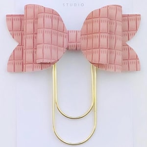 May include: A pink patterned bow with a gold double-loop paper clip. The bow is attached to a white card with the text "Duchess Gems Studio".