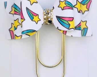 May include: A gold paper clip with a white bow decorated with a colorful shooting star pattern. The bow has a small glitter accent in the center. The text "Duchess Gems STUDIO" is printed on the white background.