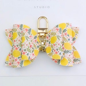 May include: A bow-shaped keychain with a lemon and floral pattern. The bow is yellow with a gold clasp and a glittery accent. The text "Duchess Gems STUDIO" is visible on the white background.