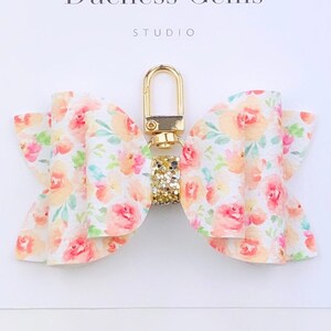 May include: A floral bow keychain with a gold-colored clasp and a small, sparkling charm. The bow features a watercolor-style pattern of peach and pink roses on a white background. The text "Duchess Gems STUDIO" is visible.