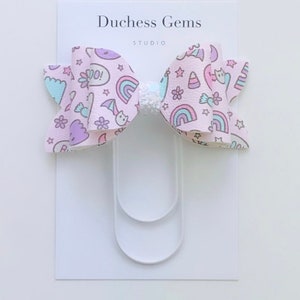 May include: A pink hair bow with a white glitter center. The bow has a pattern of rainbows, stars, and other cute designs. The bow is attached to a white alligator clip.