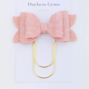 May include: A pink glitter bow hair clip with a gold-colored paper clip base. The bow is made of a textured material and is attached to a white card with the text "Duchess Gems STUDIO".