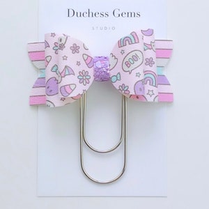 May include: A pink and purple bow with a unicorn, rainbow, and candy cane print. The bow is attached to a silver double-pronged paper clip. The bow says "Boo!" on it.