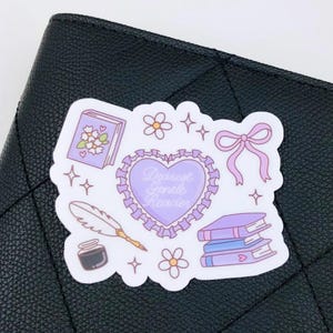 Op de afbeelding: Een sticker met een lavendel en wit ontwerp met een hart met de tekst "Dearest Gentle Reader", boeken, bloemen, een strik, een veer en inkt. De sticker ligt op een zwart, getextureerd oppervlak.