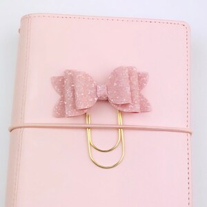 May include: A light pink planner with a matching bow-shaped paper clip. The bow is covered in glitter and attached to a gold-colored paper clip. The planner has a matching elastic band.