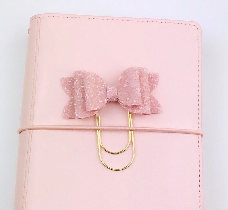 May include: A light pink planner with a matching bow-shaped paper clip. The bow is covered in glitter and attached to a gold-colored paper clip. The planner has a matching elastic band.