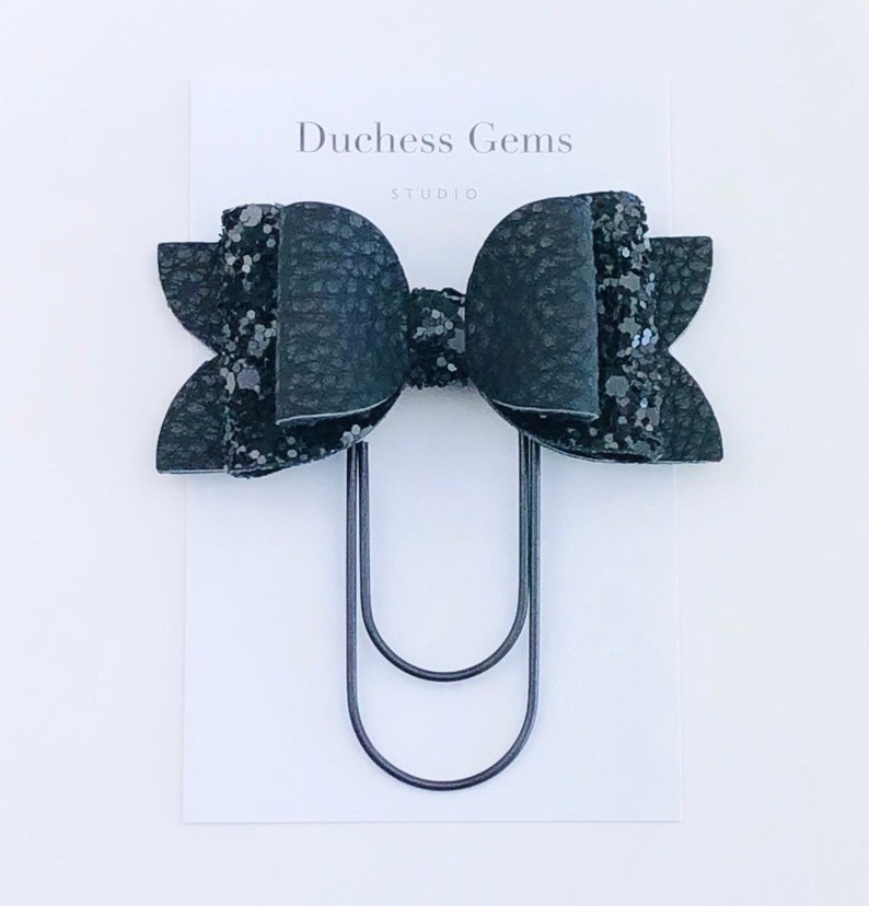 Pu&ograve; includere: Fiocco nero con glitter e una texture in similpelle nera, attaccato a una clip in metallo nero. Duchess Gems Studio.