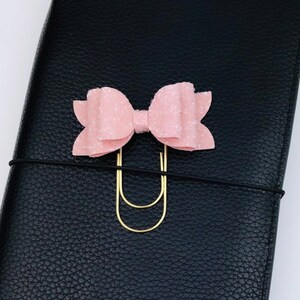 May include: A light pink bow-shaped paper clip with a gold-colored clip, attached to a black leather notebook. The bow has a textured surface and is centered on the notebook.