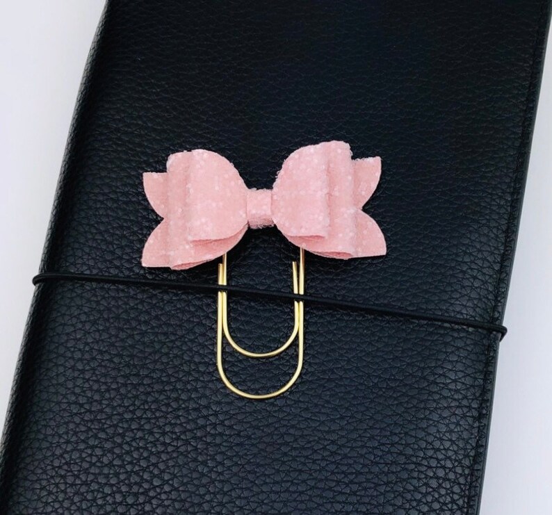 May include: A light pink bow-shaped paper clip with a gold-colored clip, attached to a black leather notebook. The bow has a textured surface and is centered on the notebook.