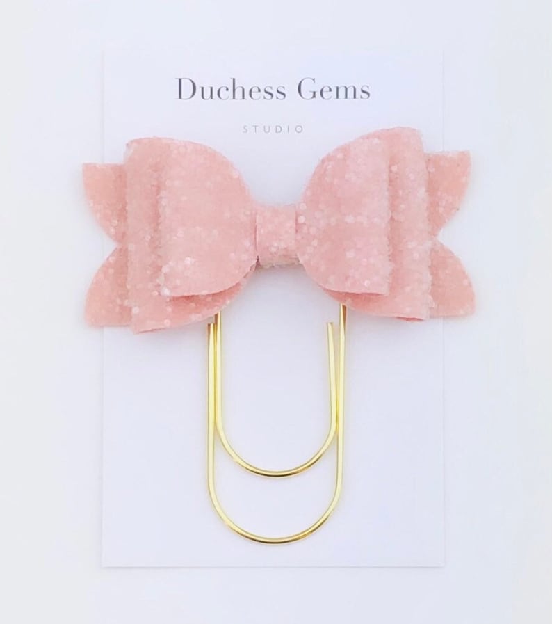 May include: A pink glitter bow hair clip with a gold-colored paper clip base. The bow is made of a textured material and is attached to a white card with the text "Duchess Gems STUDIO".