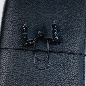 Pu&ograve; includere: Un diario rilegato in pelle nera con un fiocco di glitter nero e una clip in metallo nero.