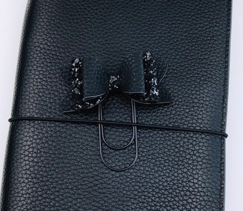 Pu&ograve; includere: Un diario rilegato in pelle nera con un fiocco di glitter nero e una clip in metallo nero.