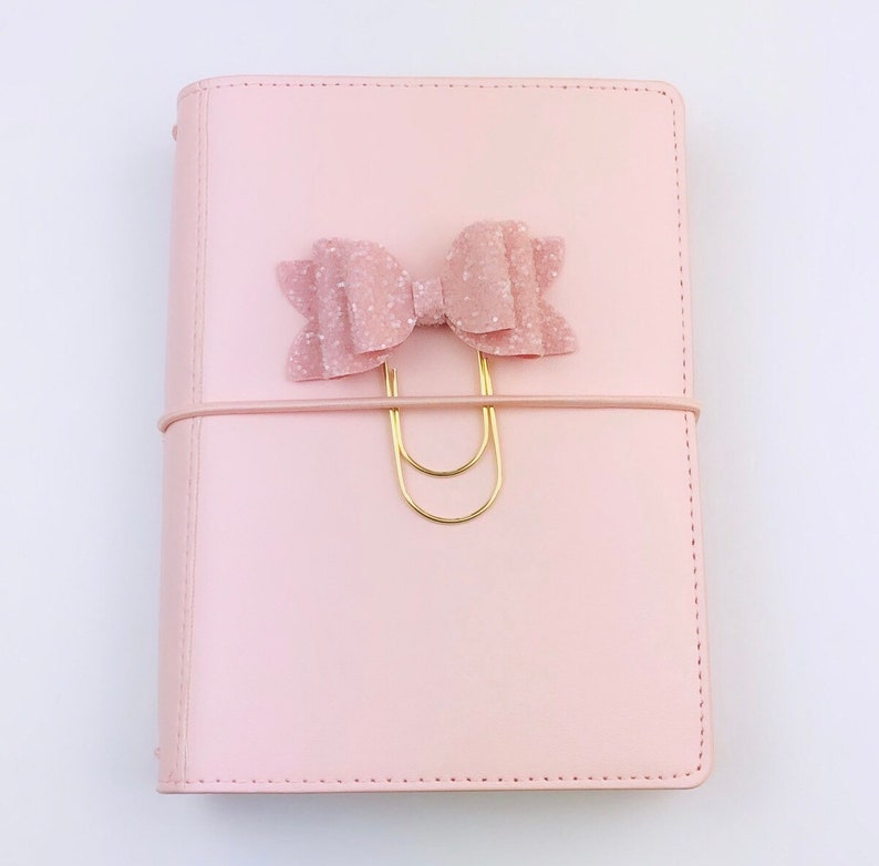 May include: A light pink planner with a matching bow-shaped paper clip. The bow is embellished with glitter. A gold-colored paper clip holds the bow in place, secured by a pink elastic band. The planner is closed.