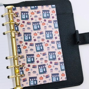 May include: A black leather-textured binder with a gold-toned six-ring mechanism holding a patterned insert. The insert features a repeating design of coffee shops, orange pumpkins, stacks of books, coffee cups, and leaves on a light pink background.