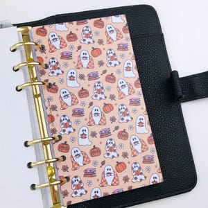 May include: A black leather six-ring binder featuring a patterned insert with a light peach background. The pattern displays white ghosts, orange pumpkins, stacks of colourful books, and small flowers in a repeating design throughout the page.