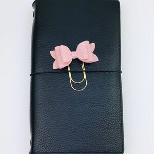 May include: Black leather journal with a textured finish. A light pink bow and gold-colored paper clip adorn the front. A black elastic band wraps around the journal. The journal is closed.