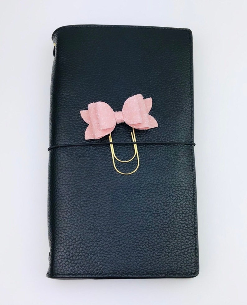 May include: Black leather journal with a textured finish. A light pink bow and gold-colored paper clip adorn the front. A black elastic band wraps around the journal. The journal is closed.