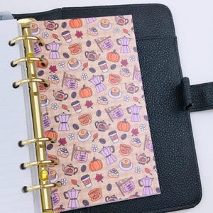 May include: A black leather-textured planner with a gold six-ring binder mechanism holding a patterned insert. The insert features a repeating design of orange pumpkins, purple coffee makers, coffee cups, and small flowers on a light tan background.