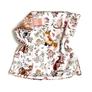 May include: A white baby blanket with a woodland animal print, including a fox, owl, deer, and rabbit. The blanket has a brown, plush border and a pink ribbon. The design features flowers, mushrooms, and butterflies.