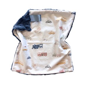 May include: A baby blanket with a cream-colored background and a pattern of various vehicles in muted colors. The blanket has a navy blue plush border and backing. A gray tag is attached to the blanket.
