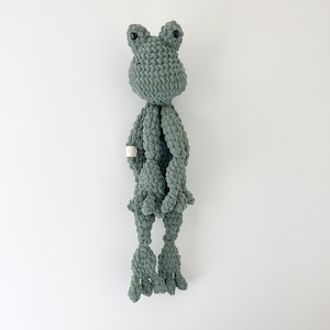 May include: A green crocheted frog toy with black eyes. The frog is standing on a white background.