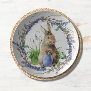 May include: A white ceramic plate with a brown bunny holding a blue egg. The bunny is surrounded by a wreath of purple flowers and green leaves.