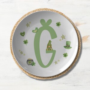 May include: A white ceramic plate with a green shamrock design. The plate features a large green letter "G" with a small gnome sitting on top of it. The plate is surrounded by a brown woven placemat.