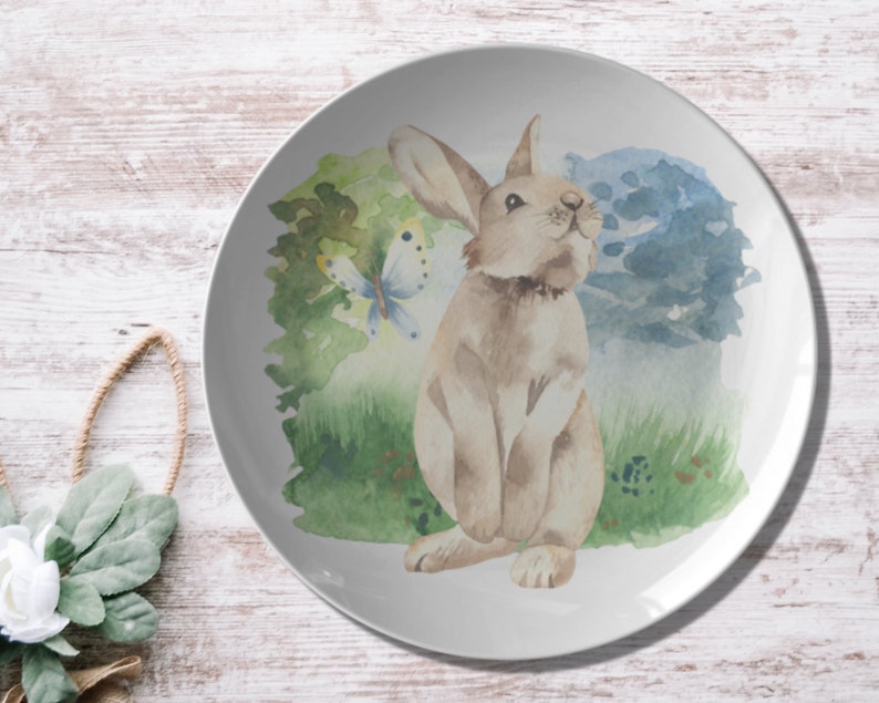 May include: A white ceramic plate with a watercolor illustration of a brown rabbit sitting in a field of green grass with a blue butterfly. The background is a watercolor wash of blue and green.