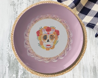 May include: A decorative plate with a Day of the Dead sugar skull design. The plate has a pink rim and a white center with a colorful skull adorned with flowers. The plate is placed on a woven placemat.