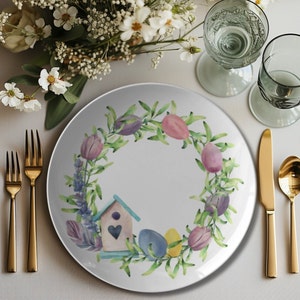 May include: A white ceramic plate with a watercolor Easter design. The plate features a wreath of green leaves, purple tulips, and pastel-colored eggs. A small birdhouse with a heart is also included. Gold cutlery and two water glasses are on the table.