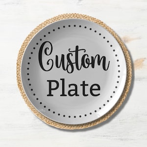 May include: A white ceramic plate with the words "Custom Plate" in black script. The plate has a dotted border and sits on a round, woven placemat. The background is a white wooden surface.