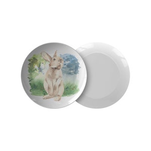 May include: A white ceramic plate with a watercolor illustration of a brown rabbit sitting in a field of green grass with a blue butterfly. The plate is part of a set of two.