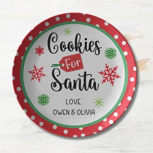 May include: A white ceramic plate with a red and green border. The plate has a white center with the text "Cookies for Santa" in black script. The text "Love. Owen & Olivia" is printed below the main text. The plate is decorated with red polka dots and green snowflakes.