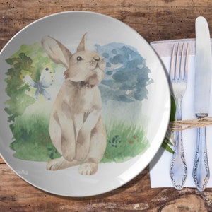 May include: A white ceramic plate with a watercolor illustration of a brown rabbit and a butterfly in a green and blue landscape.