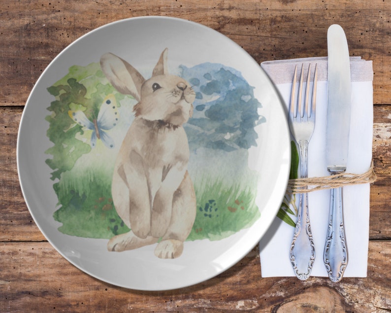May include: A white ceramic plate with a watercolor illustration of a brown rabbit and a butterfly in a green and blue landscape.