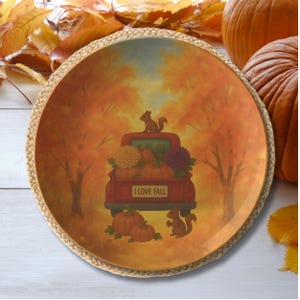May include: A round decorative plate with a fall theme. The plate features a red truck filled with pumpkins and flowers, with squirrels and the text "I LOVE FALL". The plate is surrounded by a woven border.