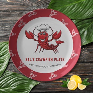 May include: Red and white round ceramic plate with a cartoon crawfish wearing a chef's hat. The plate has a white border and the text "Sal's Crawfish Plate Let the Good Times Boil".